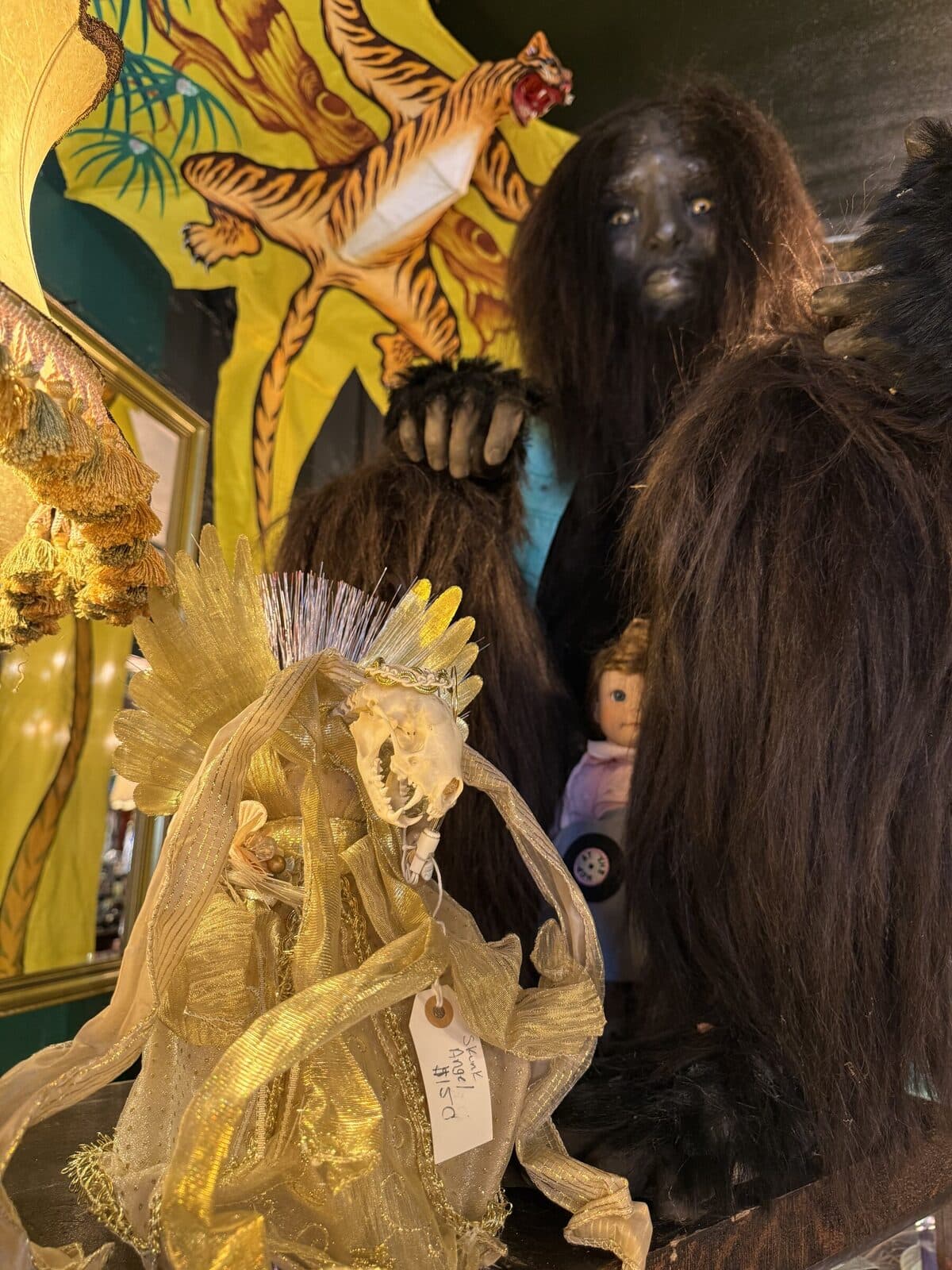 A skink-skull angel with gold feather wings, draped in metallic ribbon. A handwritten tag reads "Skink Angel $150."