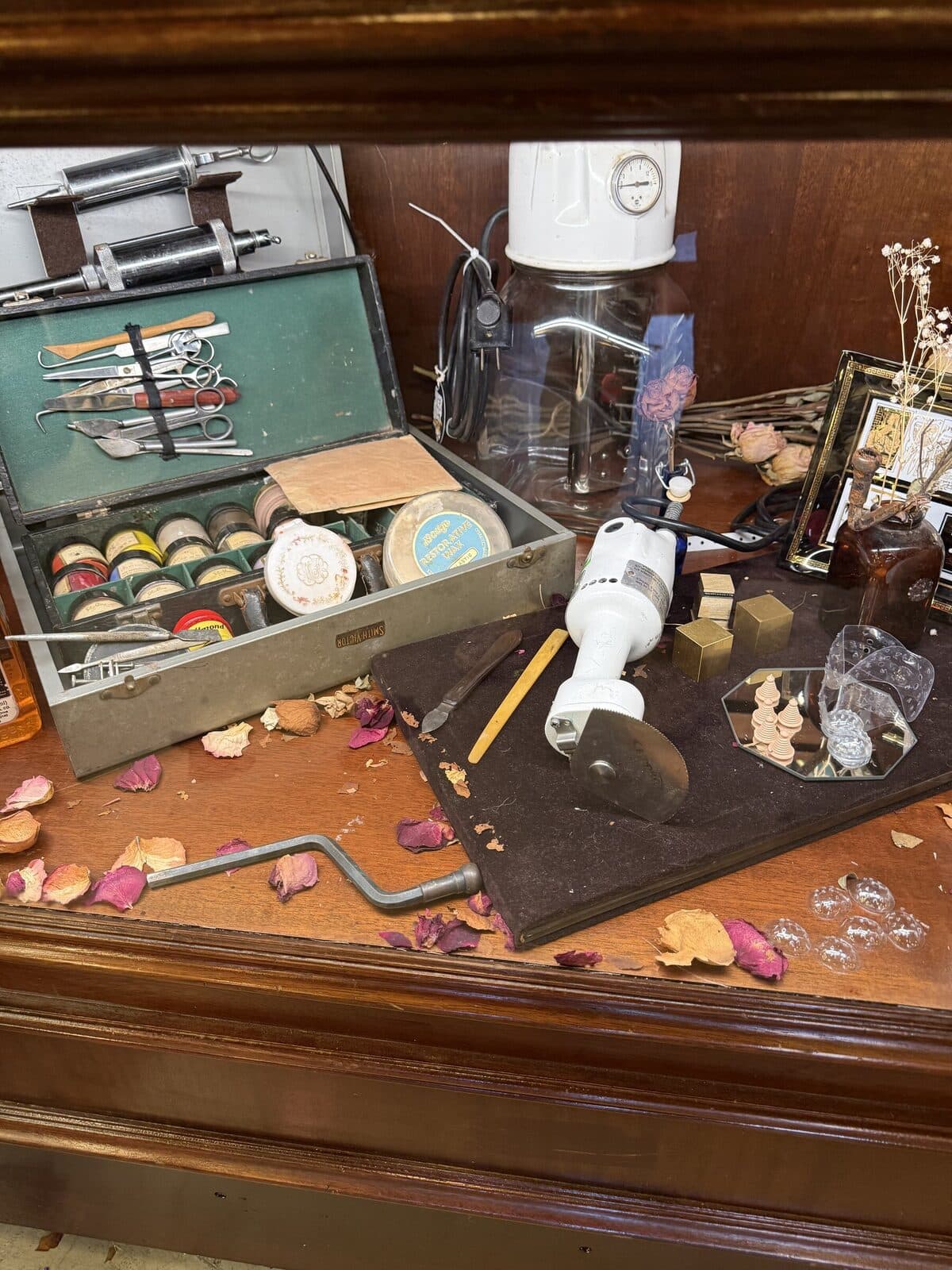A wood counter with an open vintage sewing kit (thread spools, scissors, scribe tools), a hand-cranked jeweler's tool, and a small framed photo. Dried rose petals scattered across the surface.